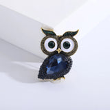 Owl brooch with dark blue faceted stone body, vintage-gold tone textured metal surface, and green accent eyes, upright on white background.
