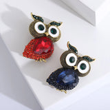 Pair of owl brooches with faceted red and blue stone bodies, textured metal detailing, and green accent eyes, angled view on white surface.