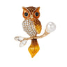 Owl brooch with gold-tone metal and orange-brown enamel finish, clear faceted stones on body, and pearl-like bead on branch, cutout on white.