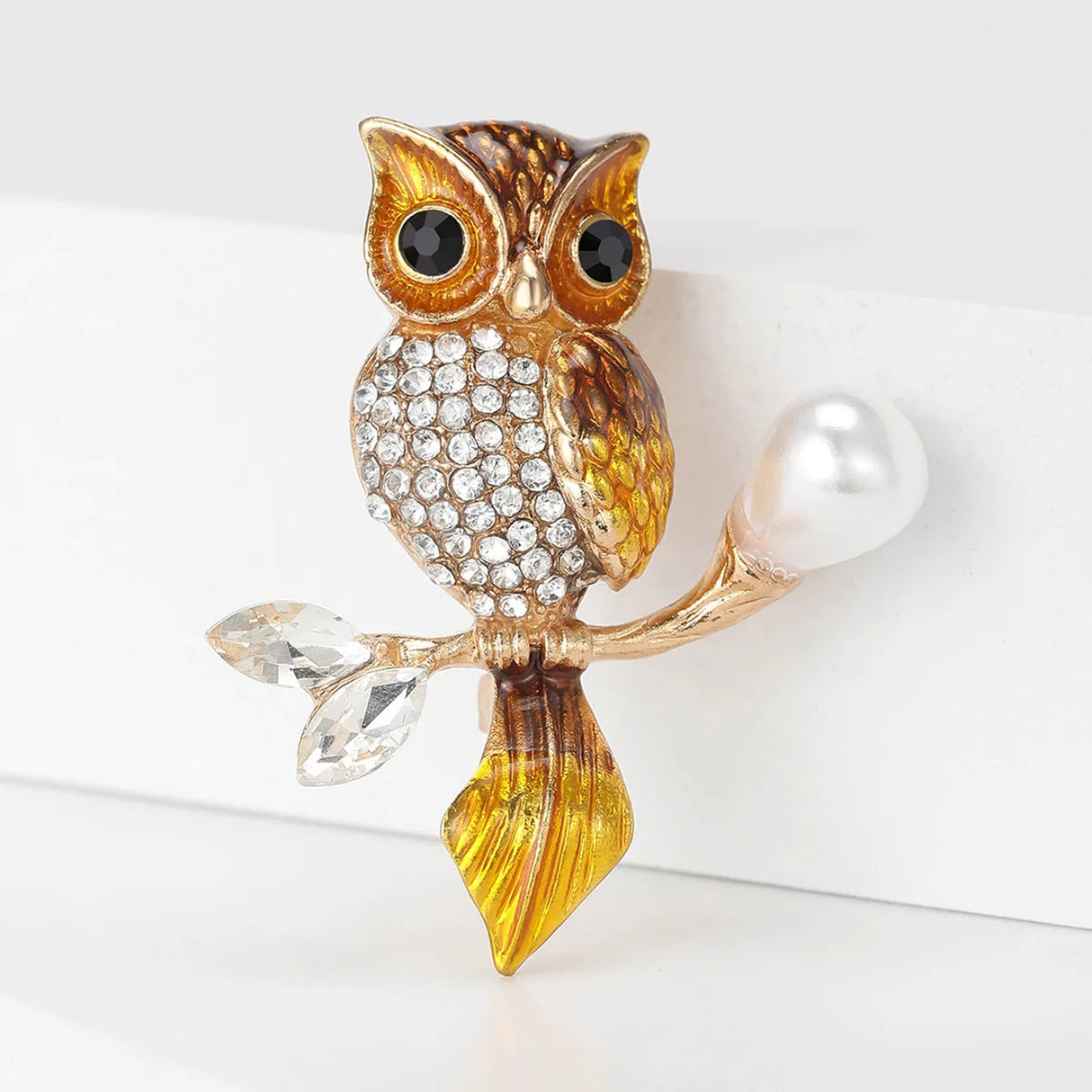 Owl brooch with gold-tone metal and brown-yellow enamel finish, clear faceted stones on the chest, and pearl-like accent on branch, angled view on white studio backdrop.