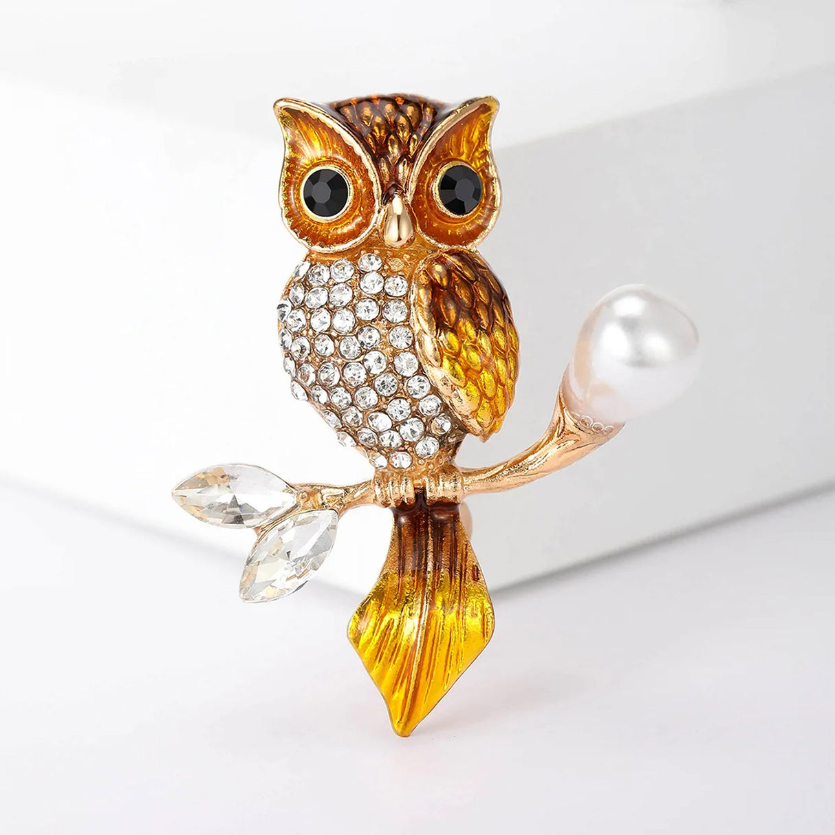 Owl brooch with orange-yellow enamel on gold-tone metal, clear faceted stones on the body, and pearl-like detail on branch, front studio view.