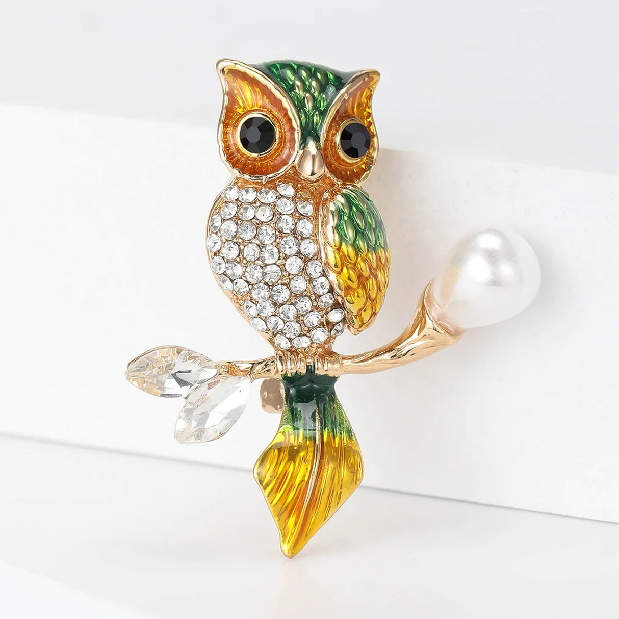 Owl brooch with green-yellow enamel and gold-tone metal finish, clear faceted stones on the chest, and pearl-like accent on branch, angled view on white.