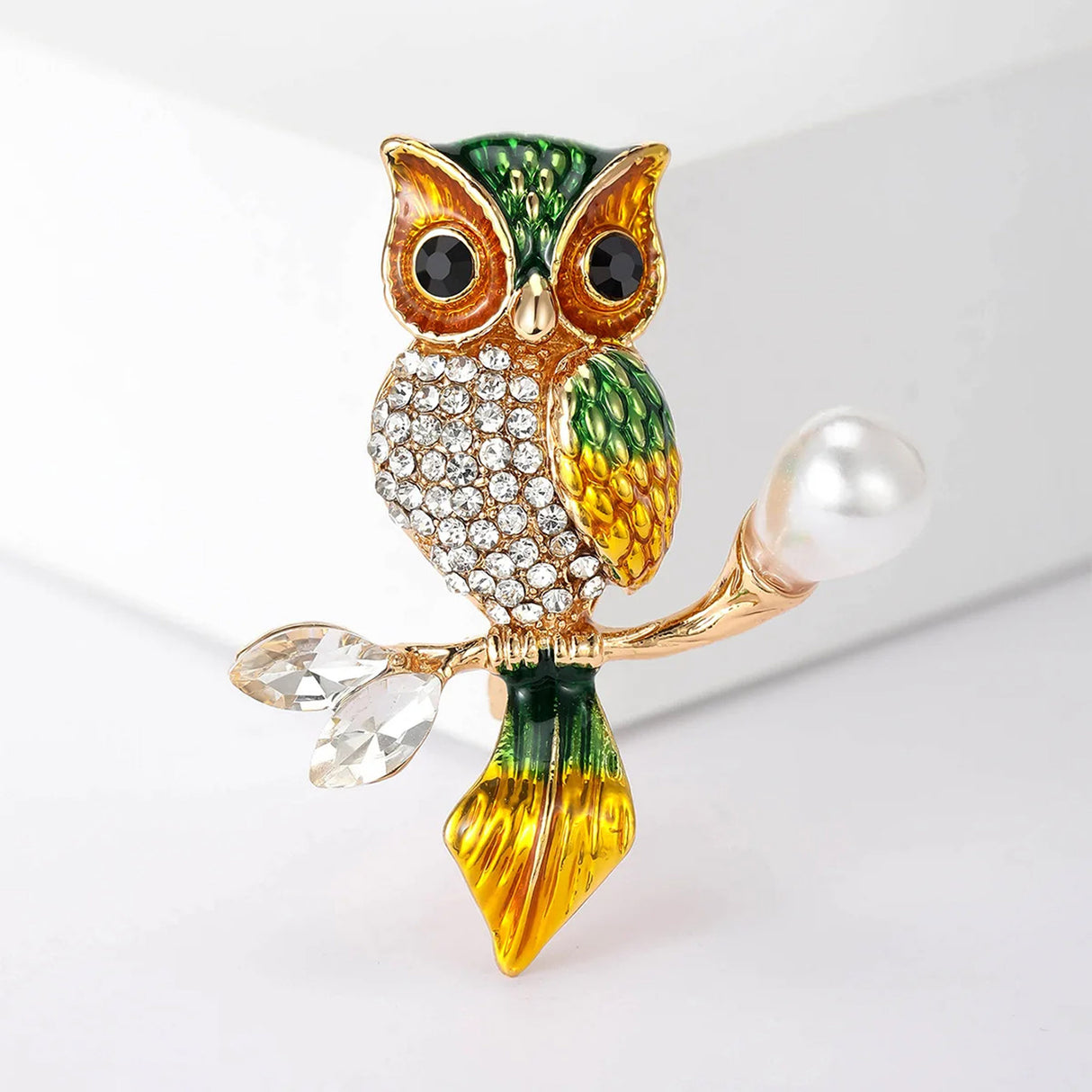 Owl brooch with green and gold-tone finish, clear faceted chest stones, and pearl-like accent on branch, front studio view on white.