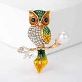 Owl brooch with green and gold-tone finish, clear faceted chest stones, and pearl-like accent on branch, front studio view on white.