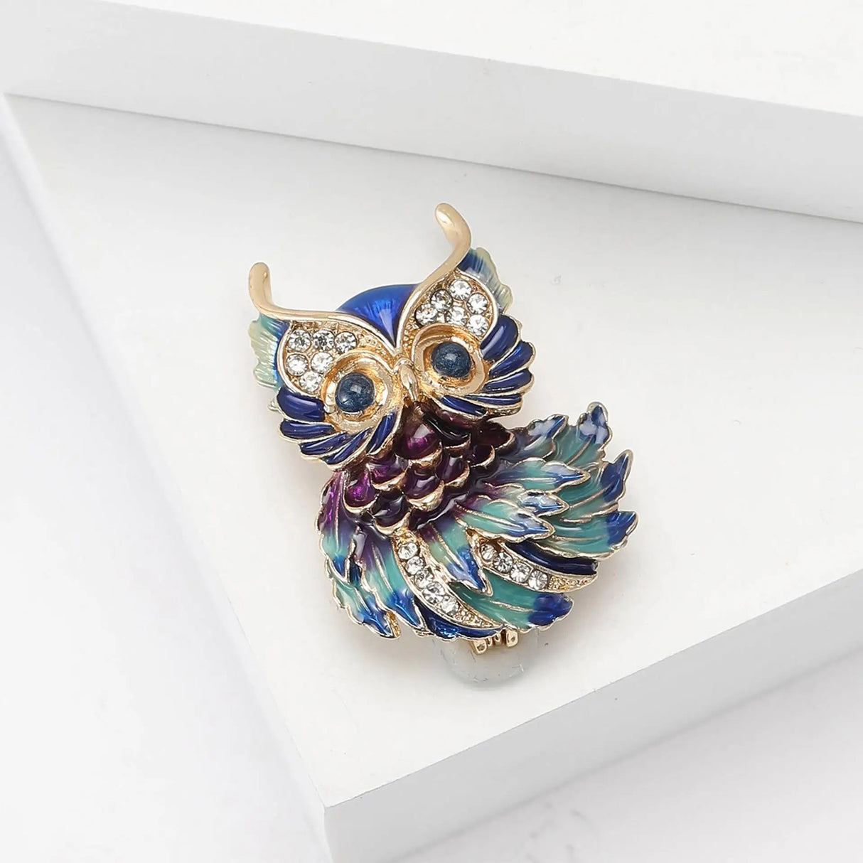 Owl brooch with gold-tone outline, blue and green enamel feathers, clear faceted stones, angled view on white block in studio.