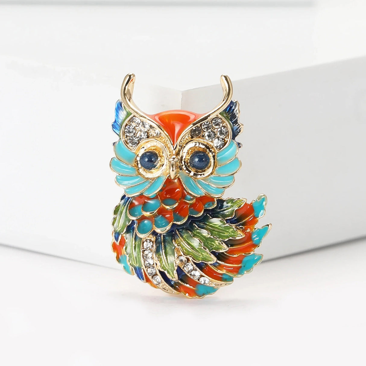 Owl brooch with gold-tone metal finish, multicolor enamel in orange, turquoise, green, and blue, clear stones, front view in studio.