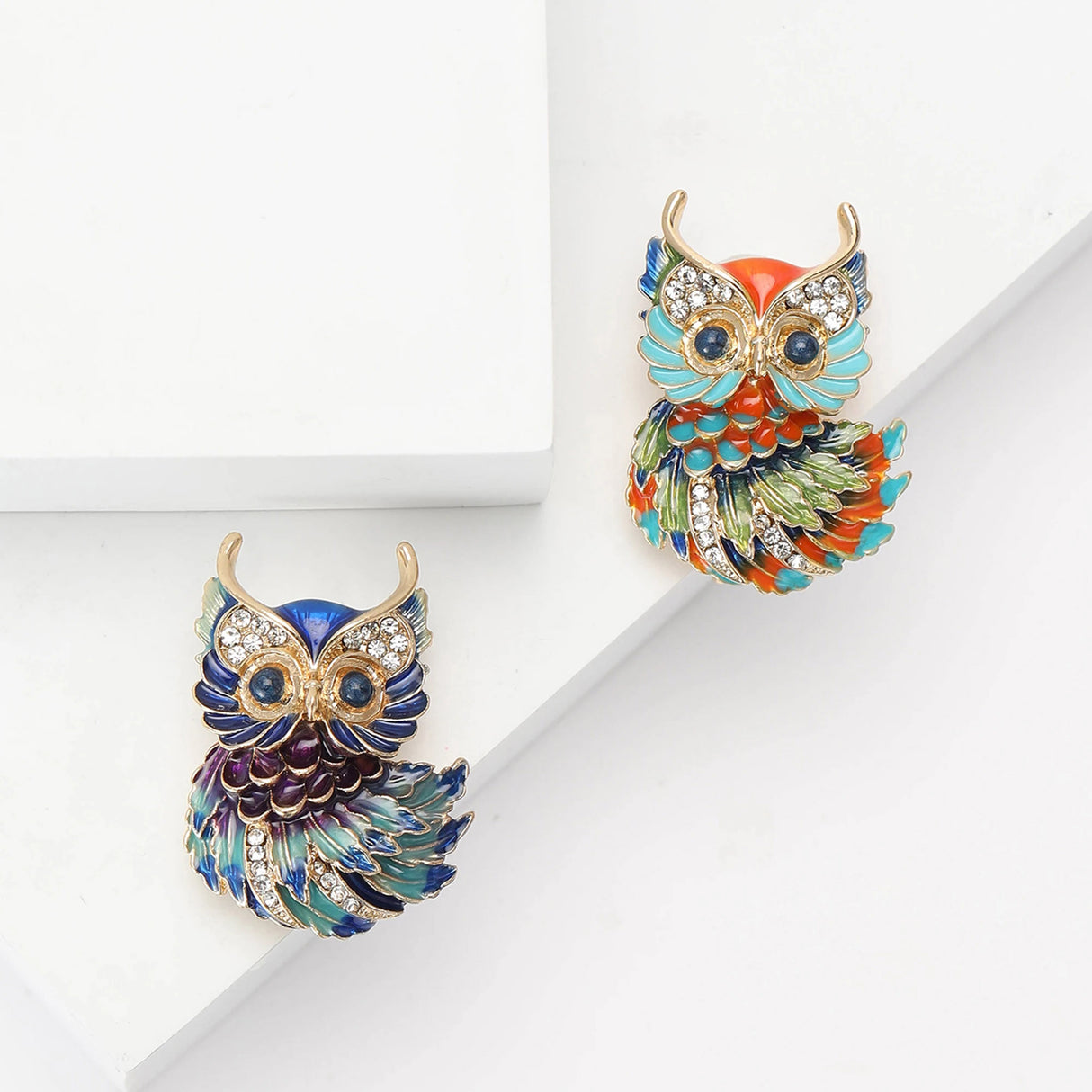 Two owl brooches with gold-tone edges, one in blue-green enamel and one in multicolor enamel, flat lay on white.