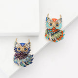Two owl brooches with gold-tone edges, one in blue-green enamel and one in multicolor enamel, flat lay on white.