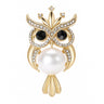 Owl brooch with gold-tone metal finish, black crystal eyes, clear round faceted stone accents, and large white round faux pearl body, cutout on white.