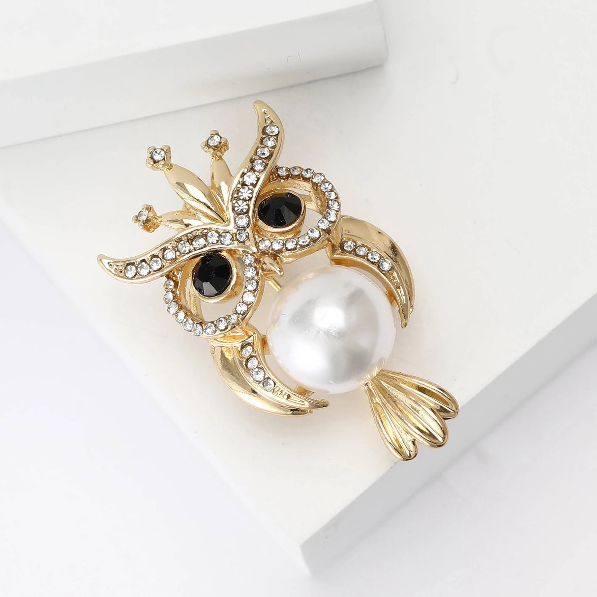 Owl brooch set in gold-tone metal with black eyes, clear rhinestone details, and round white bead body, angled view on white.
