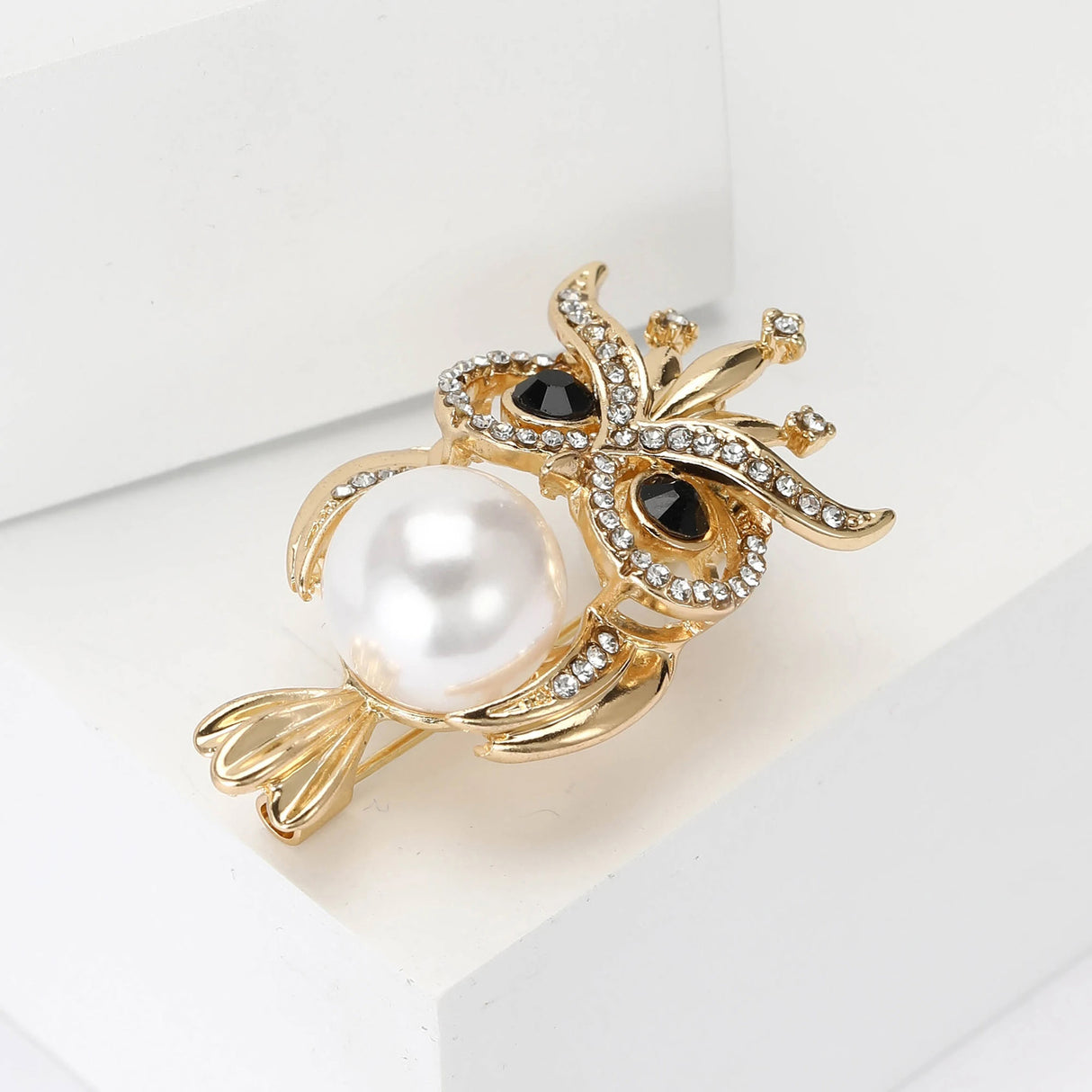 Owl brooch featuring gold-tone metal, clear faceted stones, black eye accents, and white central faux pearl, side view on white studio background.