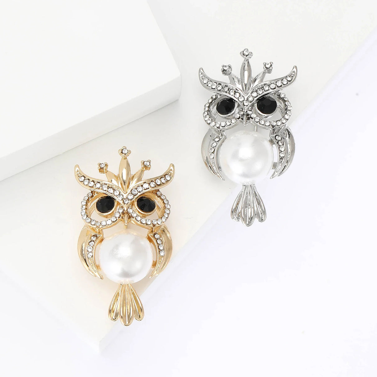 Two owl brooches in gold-tone and silver-tone metal finishes with clear stones, black eyes, and imitation pearl centers, top-down studio view.