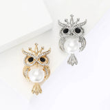 Two owl brooches in gold-tone and silver-tone metal finishes with clear stones, black eyes, and imitation pearl centers, top-down studio view.