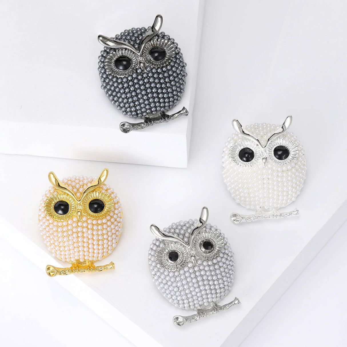 Set of four owl brooches in gray, white, gold and silver tones with small round beads and black eyes, flat lay on white blocks.