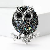 Owl brooch with black-tone mosaic body resembling abalone shell, iridescent shell-like accents, and silver-tone branch base, cutout on white.