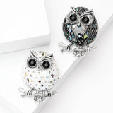 Set of two owl brooches featuring black and white-tone enamel mosaic bodies with reflective accents that resemble abalone shell, top-down view on white blocks.