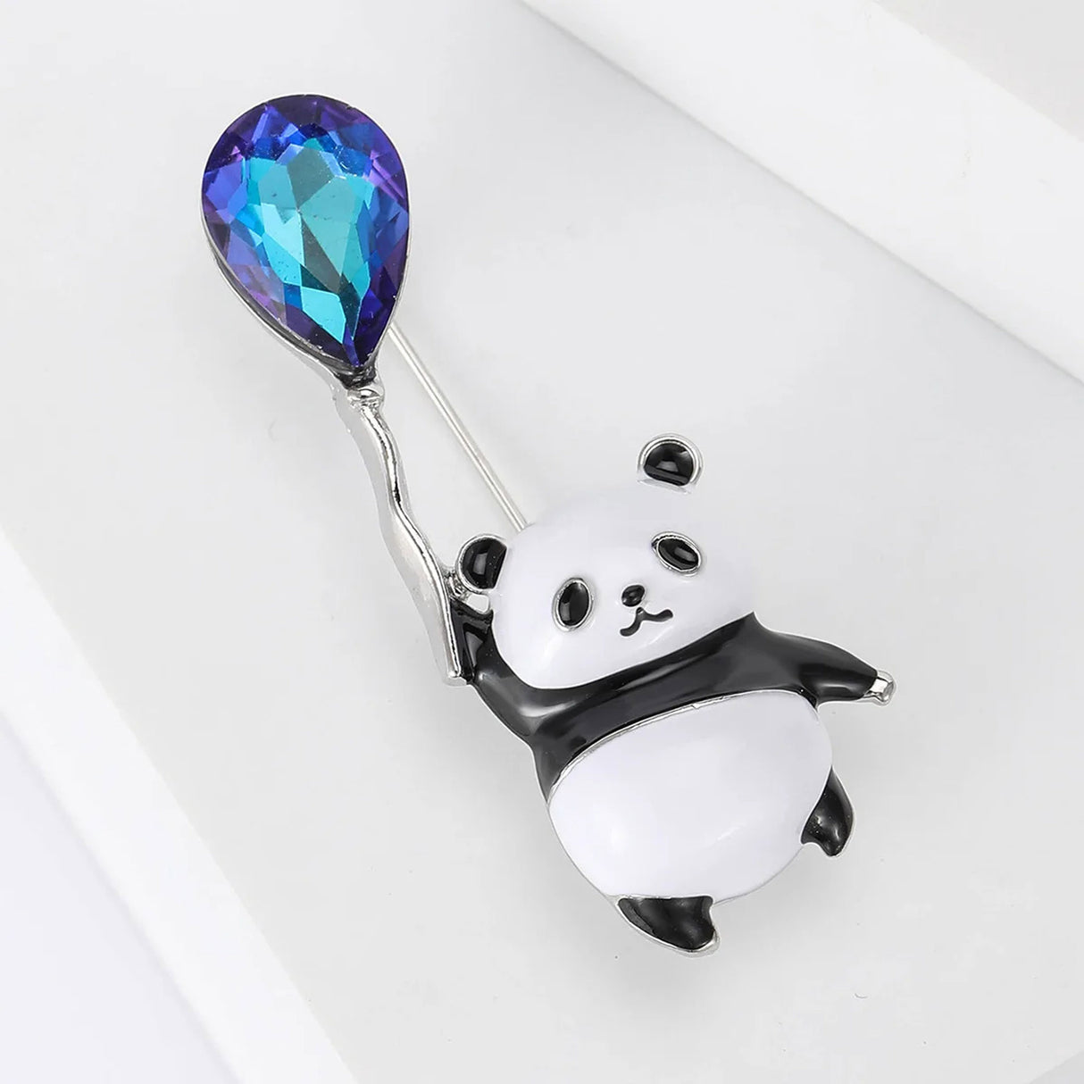 Panda brooch with black and white enamel, holding a blue and purple faceted balloon-shaped stone, angled bird's eye view on white.
