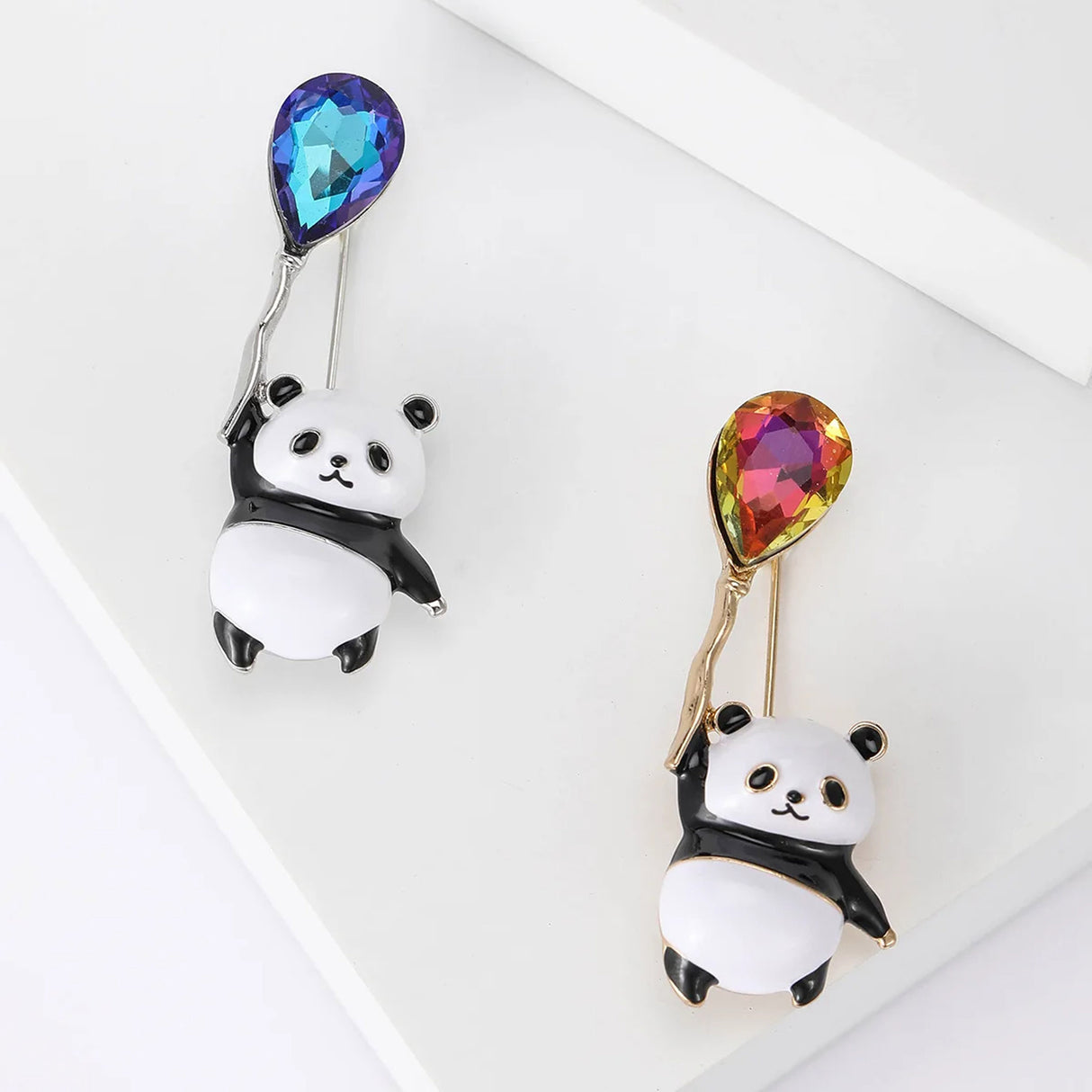 Set of two panda brooches with black and white enamel, one holding blue faceted balloon stone and one with orange-pink, flat lay on white.