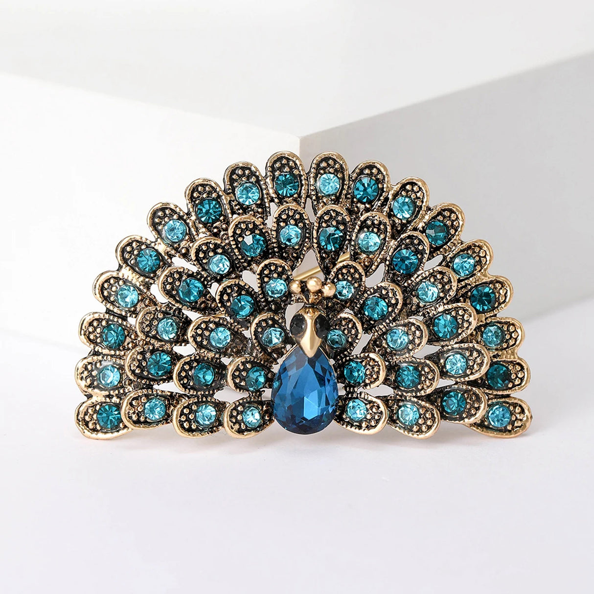 Peacock brooch with gold-tone metal, bright blue and teal stones, and a teardrop center stone, cutout on white.
