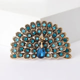 Peacock brooch with gold-tone metal, bright blue and teal stones, and a teardrop center stone, cutout on white.