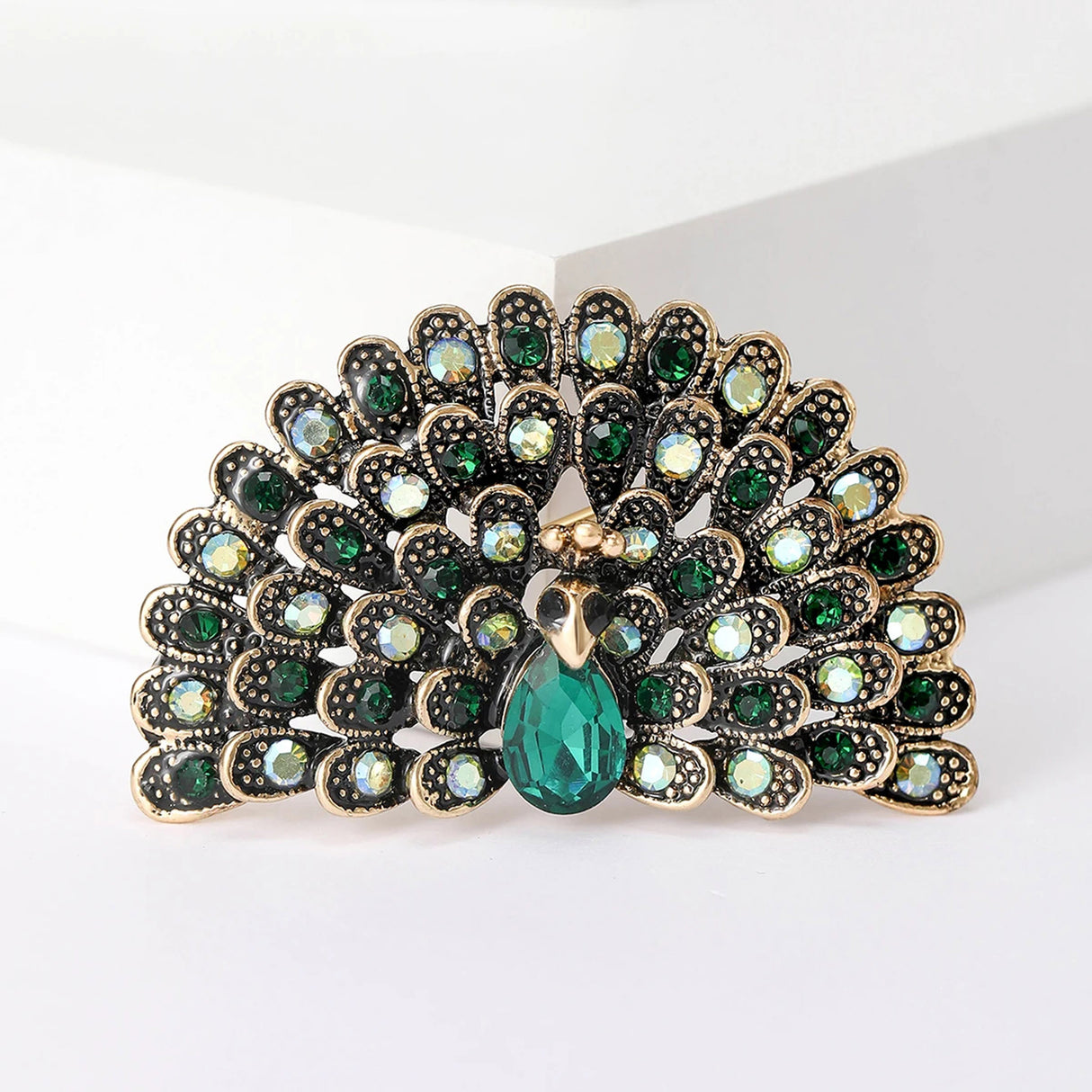 Peacock brooch with gold-tone base, green and iridescent stones, and faceted green teardrop center, cutout on white.