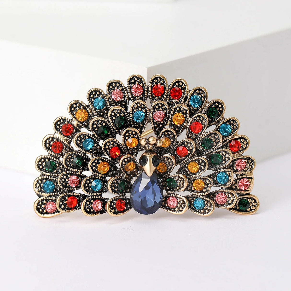 Peacock brooch in gold-tone metal with multicolor stones and faceted dark blue teardrop center, cutout on white.