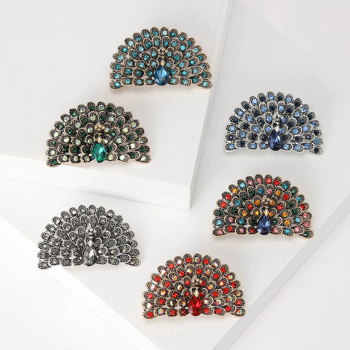 Set of six peacock brooches in assorted colors and gold- or silver-tone finishes, arranged in flat lay on white platforms.