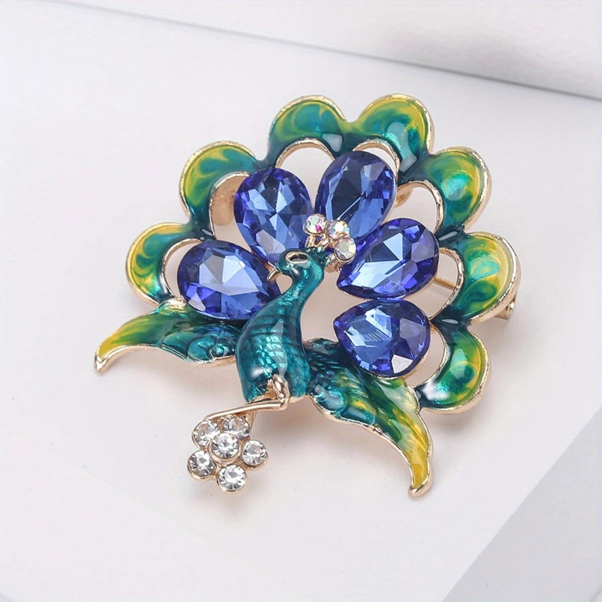 Peacock brooch with green enamel feathers, blue faceted stones, and small clear stone flower drop, angled view on white surface.