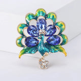 Peacock brooch with yellow-green enamel feathers, blue faceted stones, and clear crystal flower drop, upright pose on white platform.