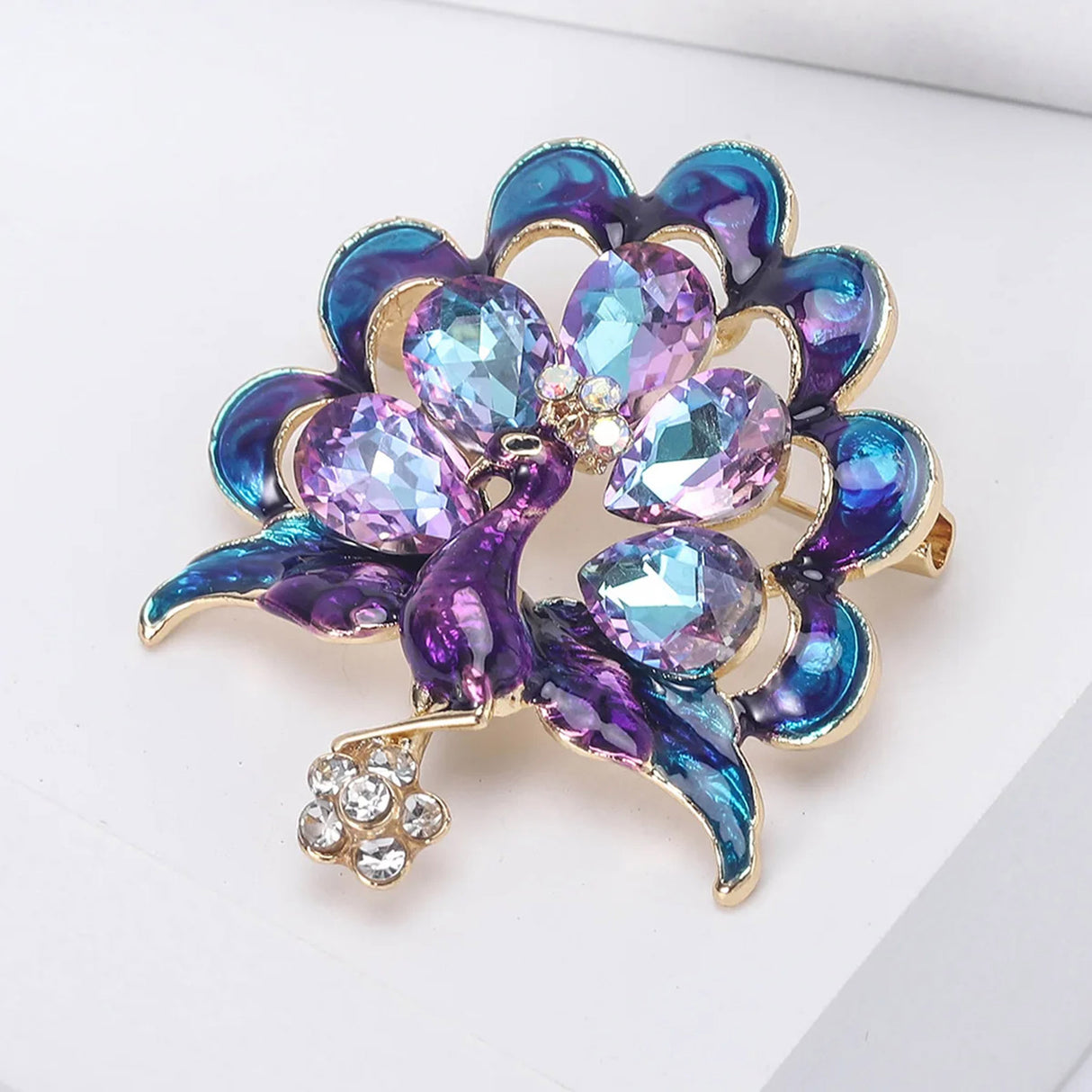Peacock brooch with metallic violet and aqua enamel feathers, lavender and icy blue faceted stones, and clear stone flower drop, angled view on white surface.
