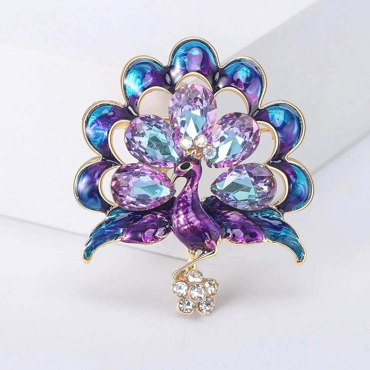 Peacock brooch with purple-blue enamel feathers and faceted stones, accented by clear stone flower drop. Upright pose on white block.
