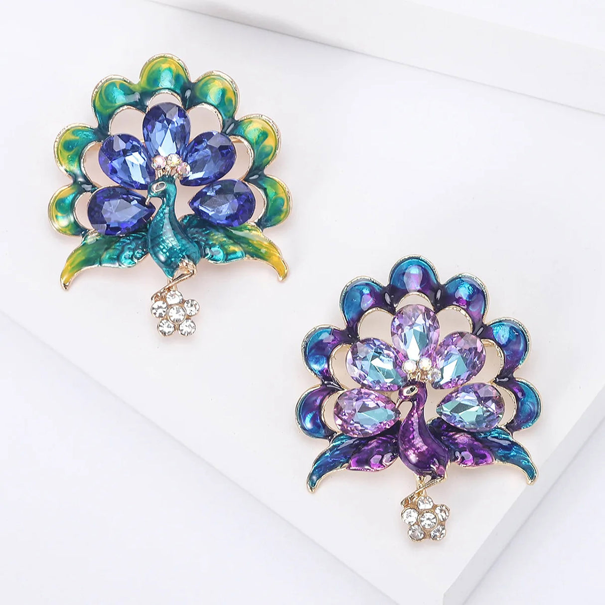Pair of peacock brooches with enamel and rhinestone feather designs. One in green-blue and one in purple-blue color combination, both featuring clear stone flower drops. Flat lay on white surface.