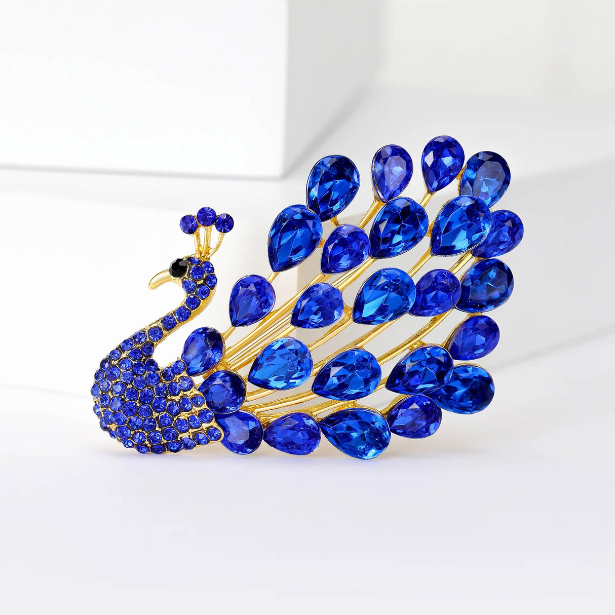 Peacock brooch with gold-tone base, blue teardrop faceted stones, and small round stones, upright on white.