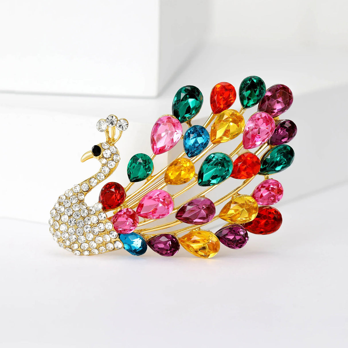 Peacock brooch with tail made from multicolor teardrop faceted stones and gold-tone finish, upright pose on white background.