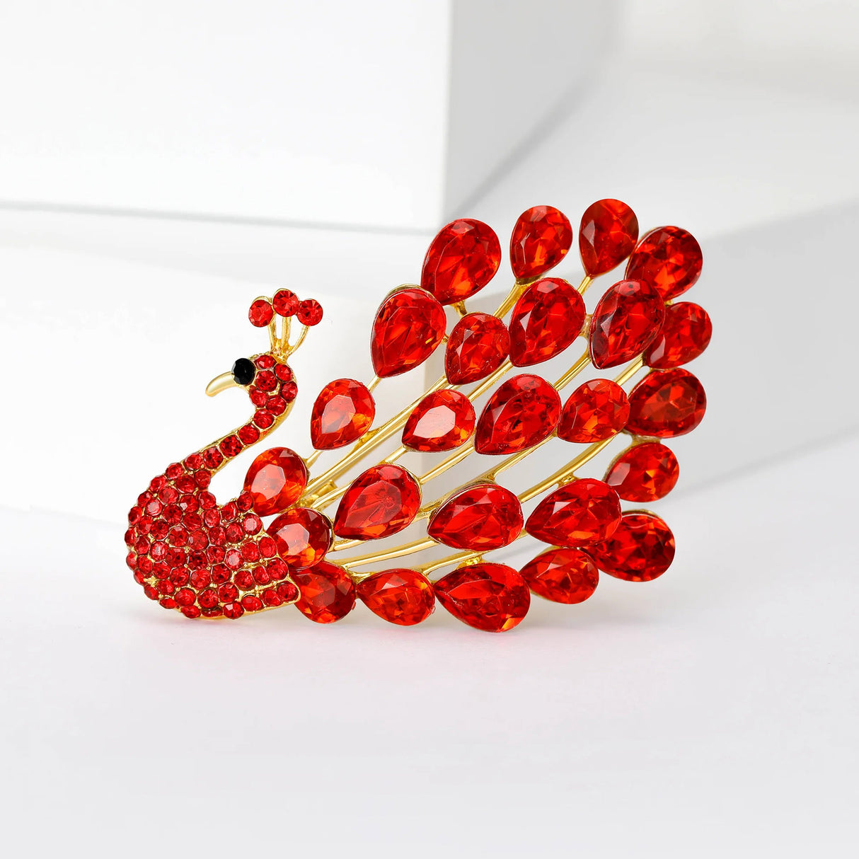 Peacock brooch with red teardrop faceted stones and small matching round stones on a gold-tone base, upright view on white.