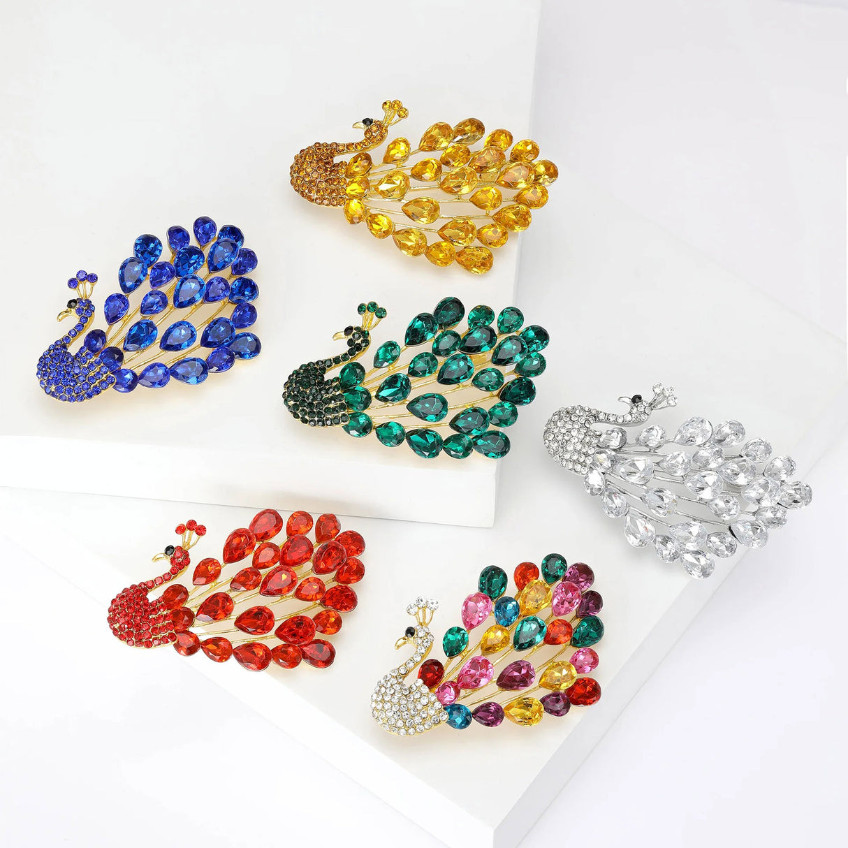 Set of six peacock brooches in multicolor, red, blue, green, yellow, and white variants, flat lay on white blocks.