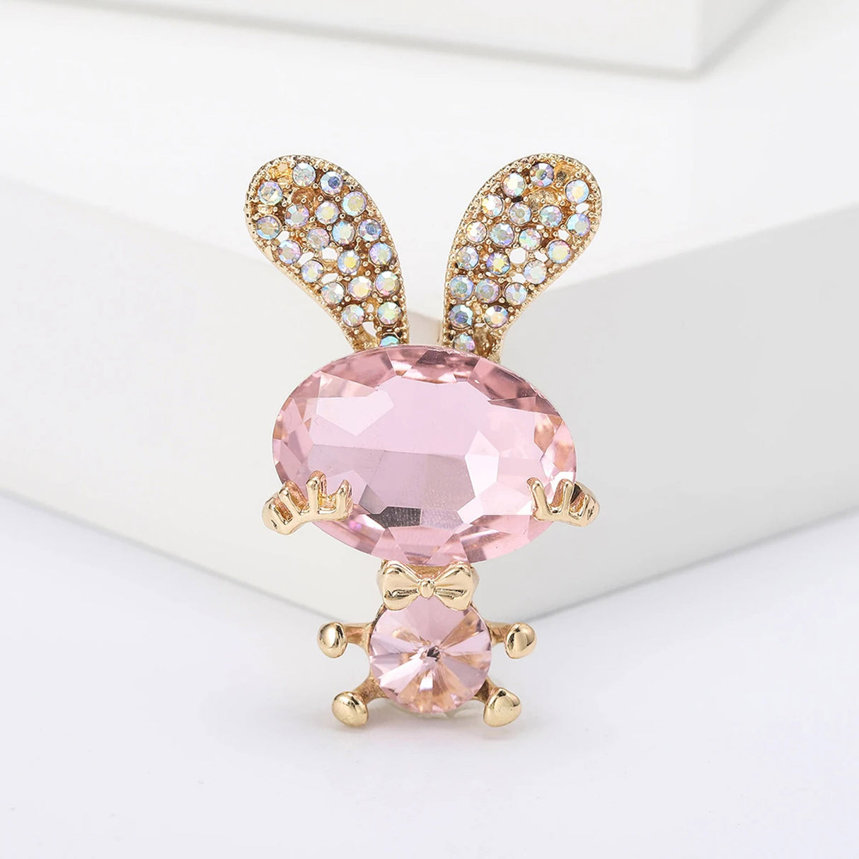 Bunny brooch with pink faceted stones, gold-tone arms and legs, and decorative crystal ears, upright pose on white block in studio.