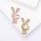 Pair of bunny brooches with pink and beige-amber faceted stones, gold limbs, and crystal-encrusted ears, flat lay on white.