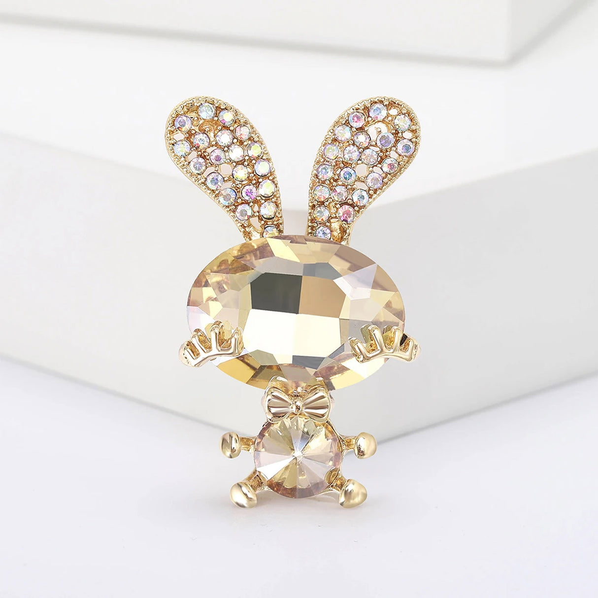 Bunny brooch featuring beige-yellow faceted stones, gold-tone limbs and bow, and crystal-studded ears, upright display on white block in studio.