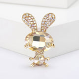 Bunny brooch featuring beige-yellow faceted stones, gold-tone limbs and bow, and crystal-studded ears, upright display on white block in studio.