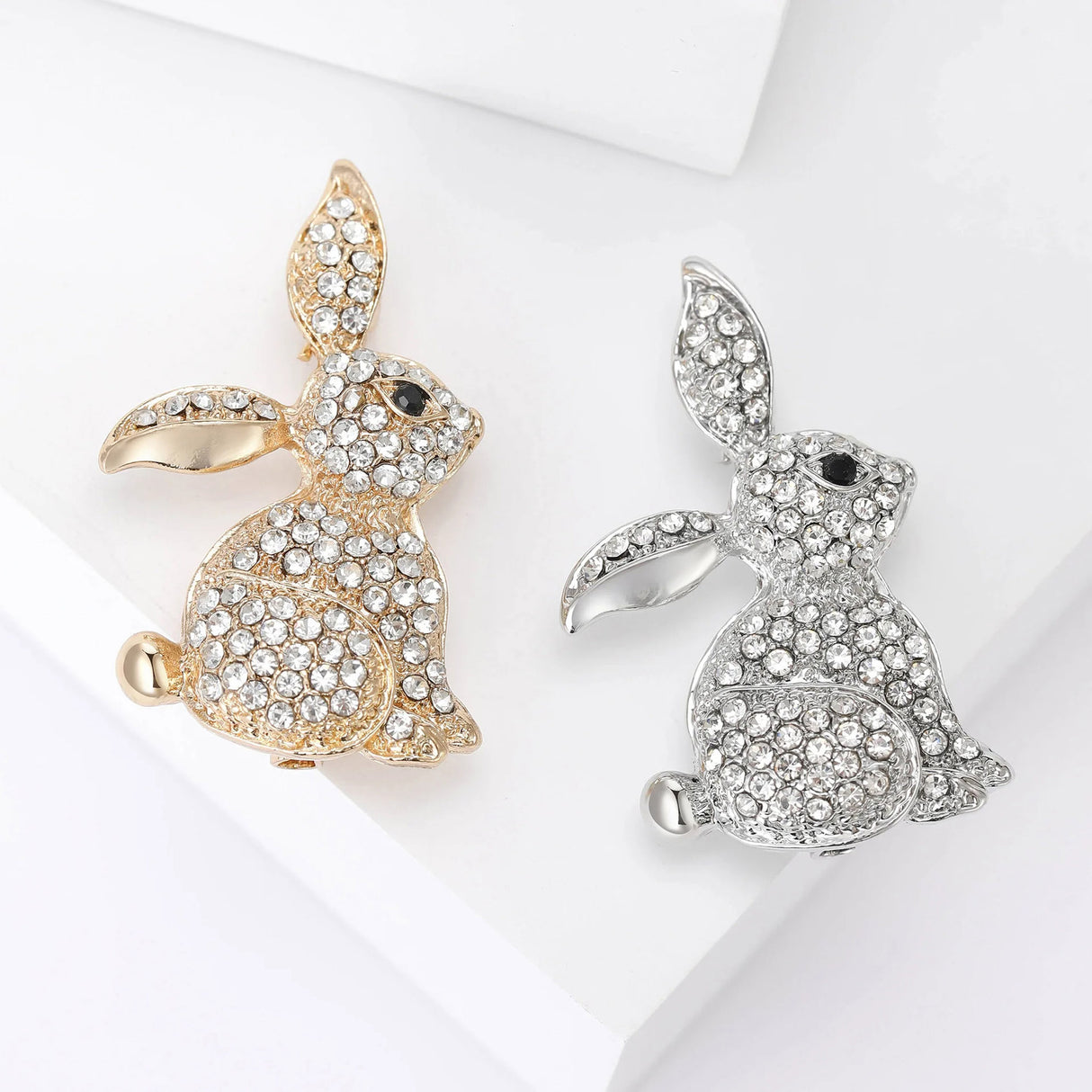 Pair of bunny brooches in gold-tone and silver-tone finishes, both with clear faceted stones and black eyes, flat lay on white surface.