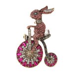 Bunny brooch with pink rhinestone pave, riding a bicycle with clear and magenta stone wheels. Cutout on white background.