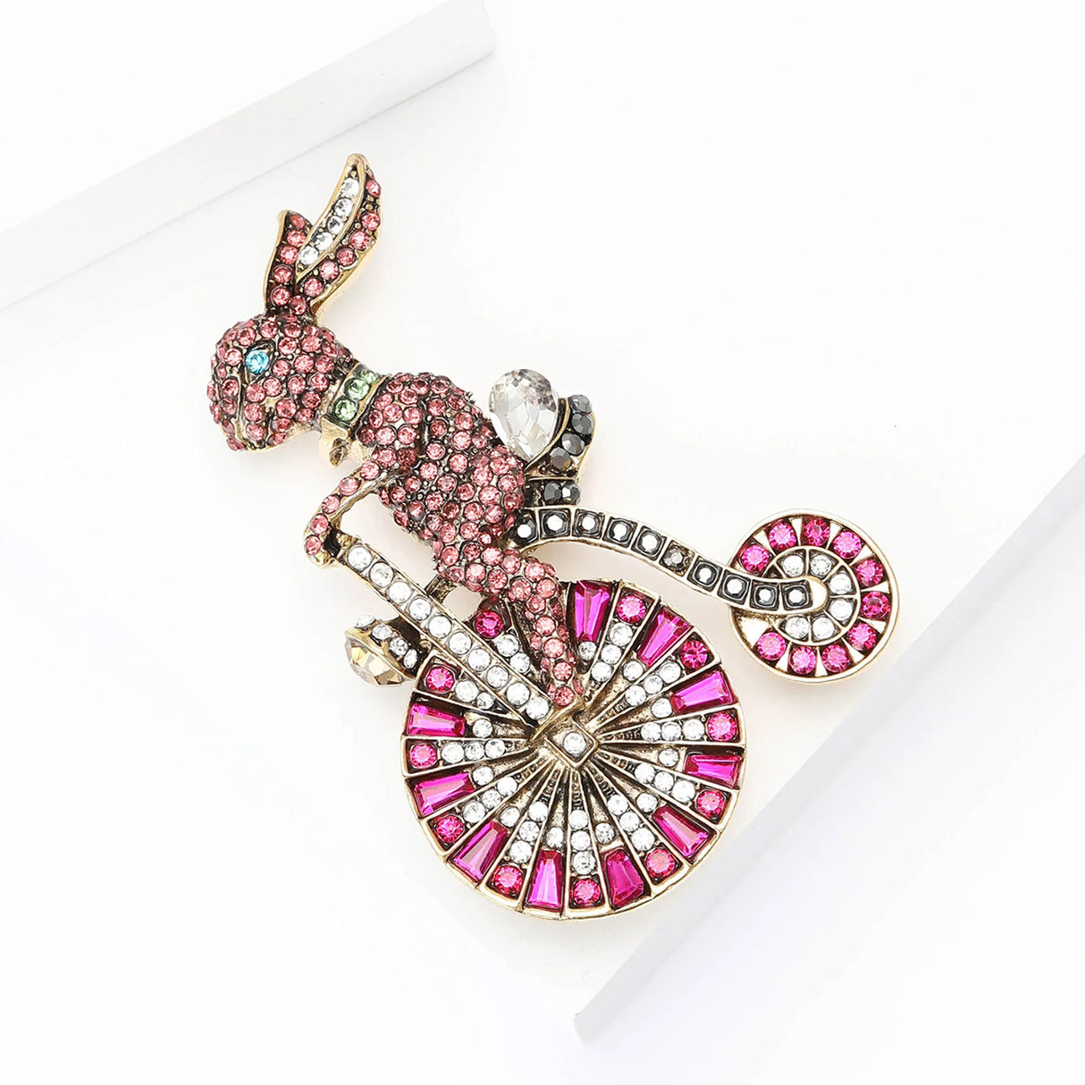 Bunny brooch with pink and white stones and blue eye detail, on bicycle with magenta and clear stone wheels, angled top view on white studio platform.
