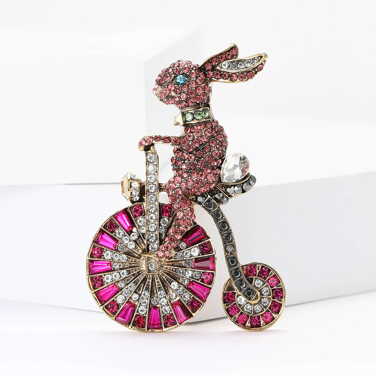 Bunny brooch covered in small round pink faceted stones, and magenta-accented bicycle wheels. Front view on white block in studio.