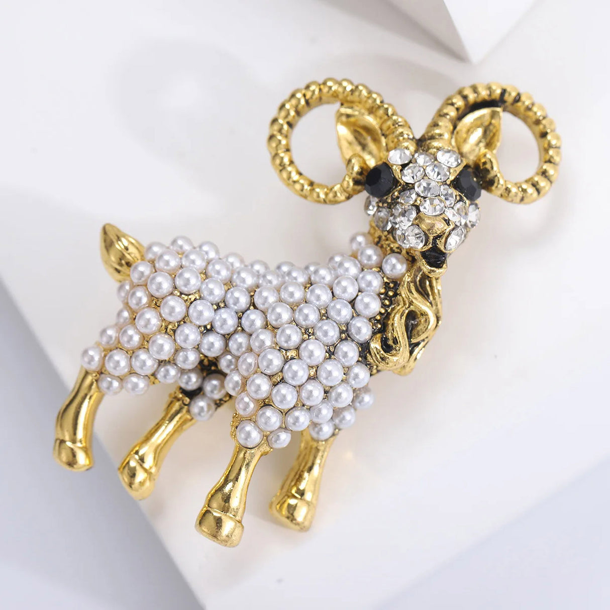 Ram brooch with gold-tone finish, white round bead body, and clear faceted stones on the face, angled side view.