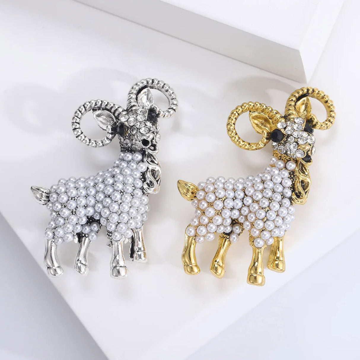 Pair of ram brooches in gold-tone and silver-tone metal finishes, with white imitation pearl bodies and faceted stone heads. Flat lay on white.