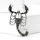 Scorpion brooch with silver-tone base, black faceted stones, and open claw design, upright pose on white block.