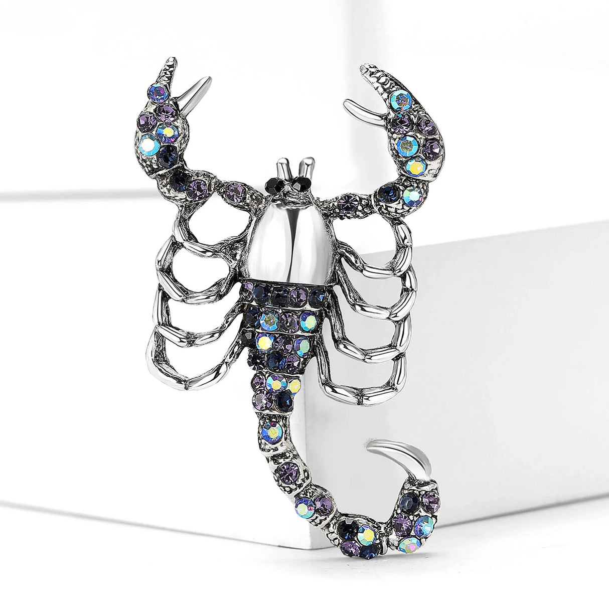Scorpion brooch with silver-tone base, iridescent blue-purple stones, and curled tail, upright view on white block.