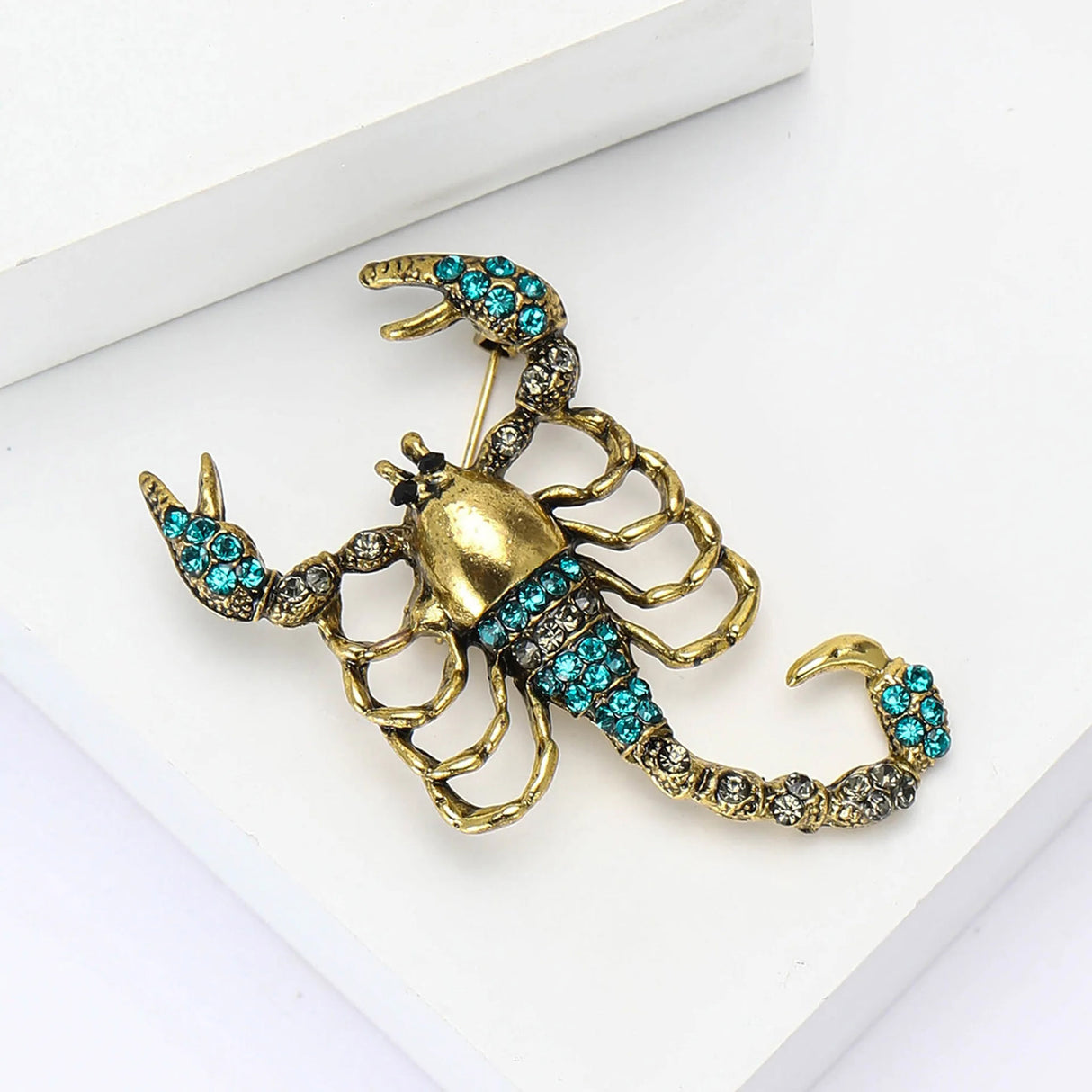 Scorpion brooch with gold-tone base, teal and clear stones, angled flat lay on white surface.