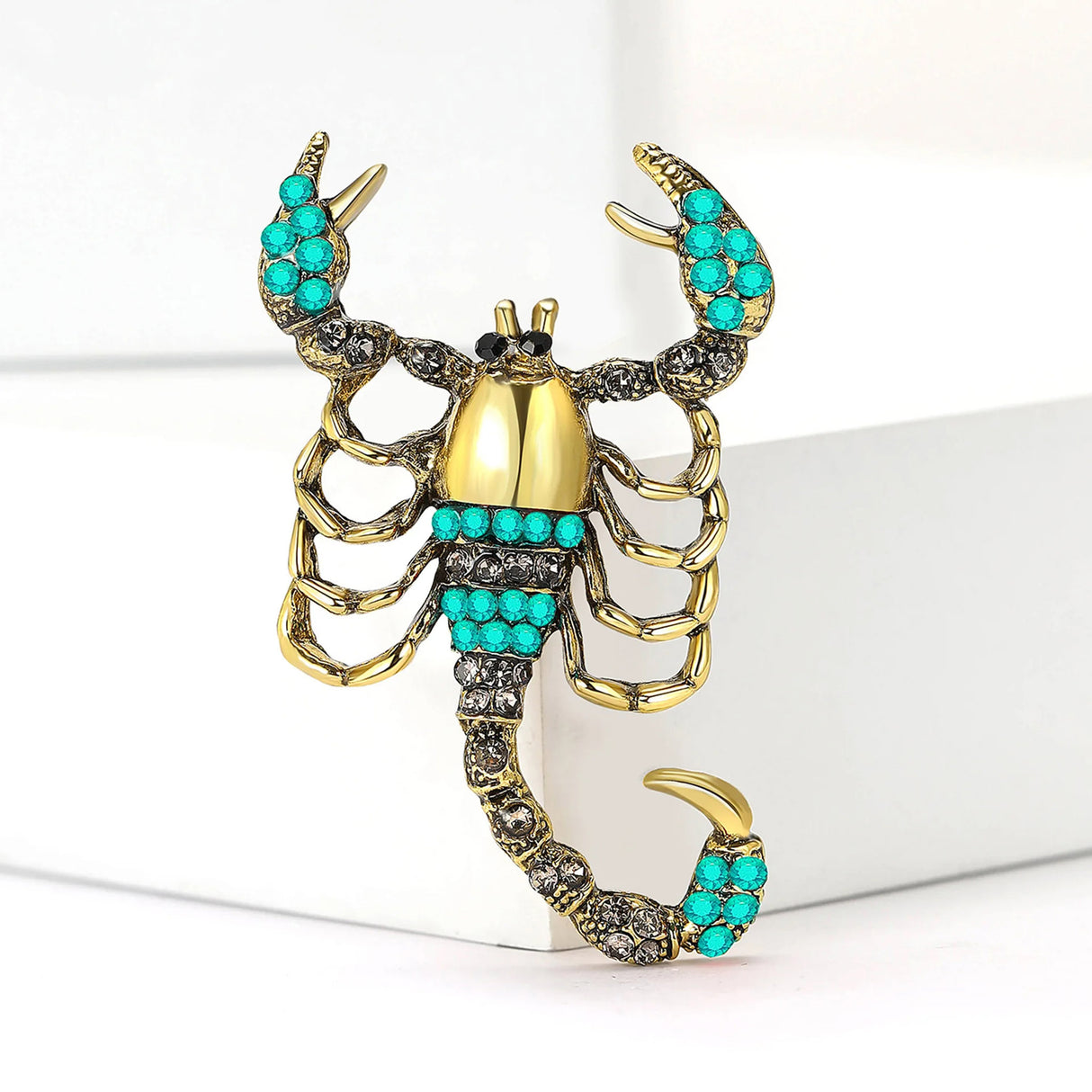 Scorpion brooch with gold-tone finish, teal and clear decorative stones, upright pose on white block.